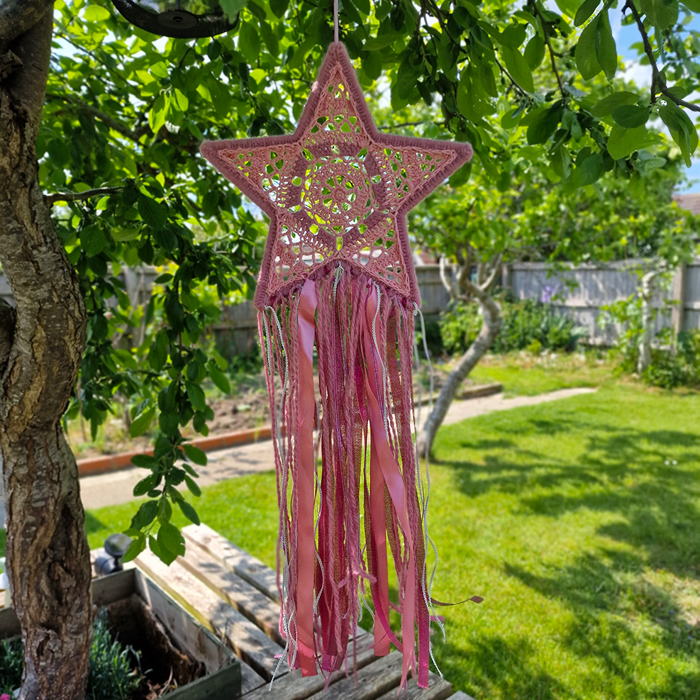 Handmade crochet star dreamcatcher with pink and silver ribbons and fringe, perfect for boho wall décor and nursery spaces.