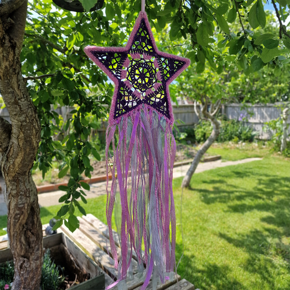 Handmade crochet star dreamcatcher with purple and pink ribbons and fringe, perfect for boho wall décor and nursery spaces.