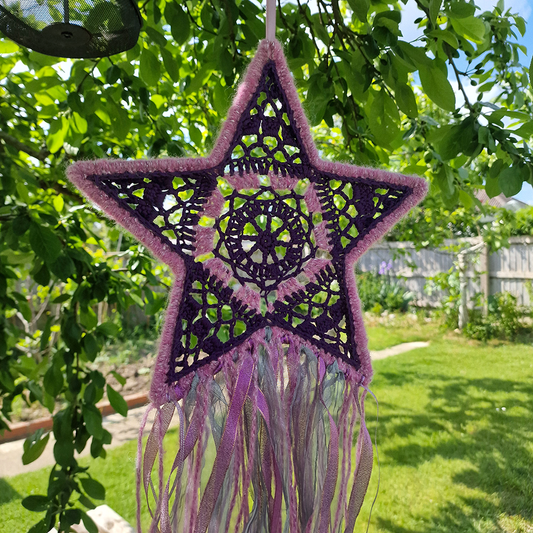 Handmade crochet star dreamcatcher with purple and pink ribbons and fringe, perfect for boho wall décor and nursery spaces.