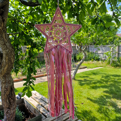 Handmade crochet star dreamcatcher with pink and silver ribbons and fringe, perfect for boho wall décor and nursery spaces.