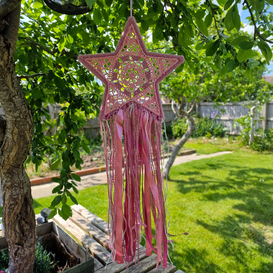 Handmade crochet star dreamcatcher with pink and silver ribbons and fringe, perfect for boho wall décor and nursery spaces.