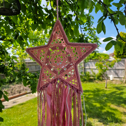 Handmade crochet star dreamcatcher with pink and silver ribbons and fringe, perfect for boho wall décor and nursery spaces.